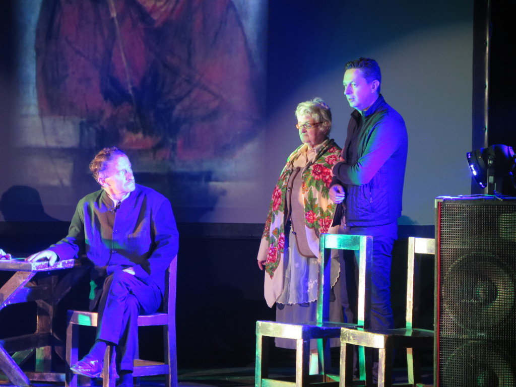 Zdjęcie ze spektaklu Brat naszego Boga w Proszowicach. Aktorzy grają na scenie w tle obraz ecce homo adama chmielowskiego. Mężczyzna przy stole siedzi i patrzy na starszą kobietę, którą trzyma pod ramię mężczyzna w średnim wieku.