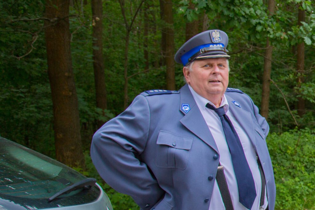 Andrzej w kostiumie policjanta, znany z pierwszego sezonu komendant. W tle lasek, drzewa liściaste.