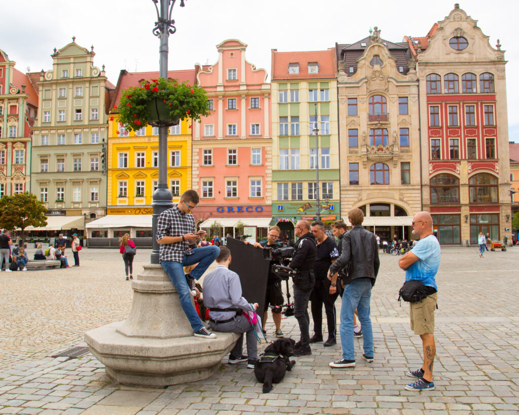 Rynek Główny we Wrocławiu; w tle kolorowe kamienice, restauracje, spacerujący ludzie. W centrum zdjęcia grupa dziewięciu osób; troje aktorów na kamiennej ławce otaczającej lampę oraz sześcioro filmowców skupionych dookoła kamery telewizyjnej. Aktor Michał, stojący na ławce, patrzy w telefon. Siedzący aktorzy Renata i Wito rozmawiają ze sobą, filmowcy także rozmawiają w grupie.