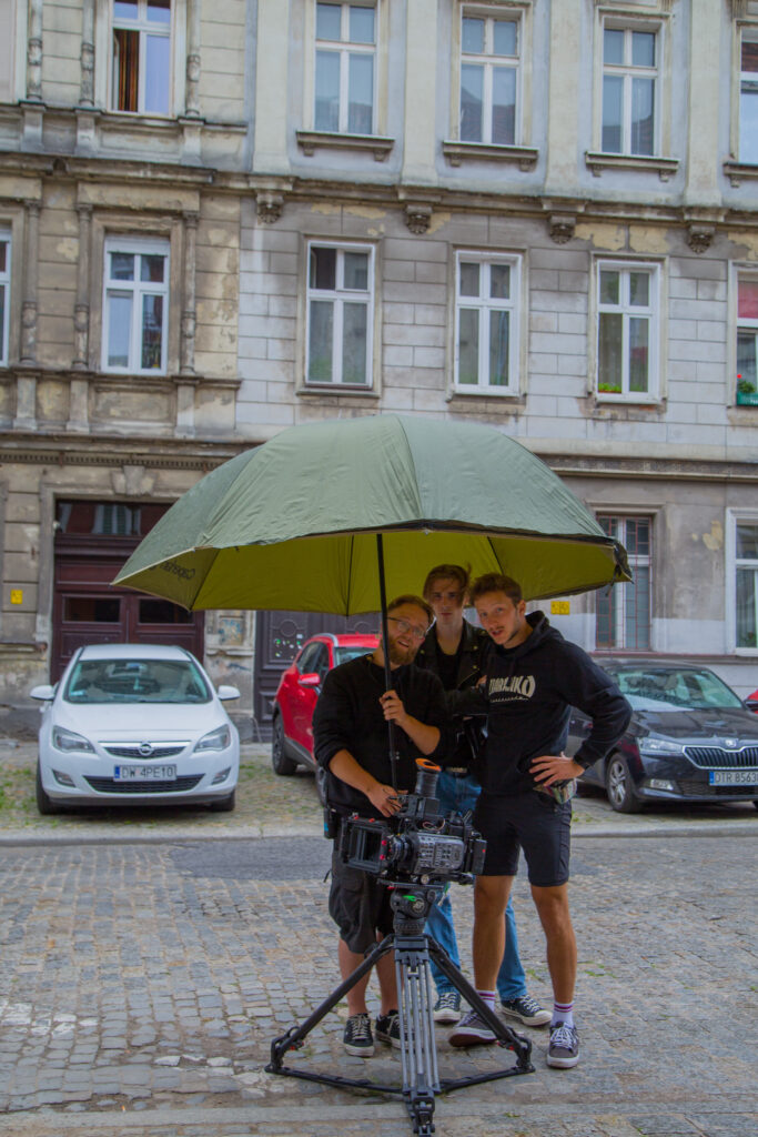 Ulica; w tle kamienica, zaparkowane samochody. W centrum zdjęcia trzech filmowców pod dużym, zielonym parasolem. Patrzą ponad obiektywem i rozmawiają. Przed nimi na bardzo niskim statywie stoi kamera .