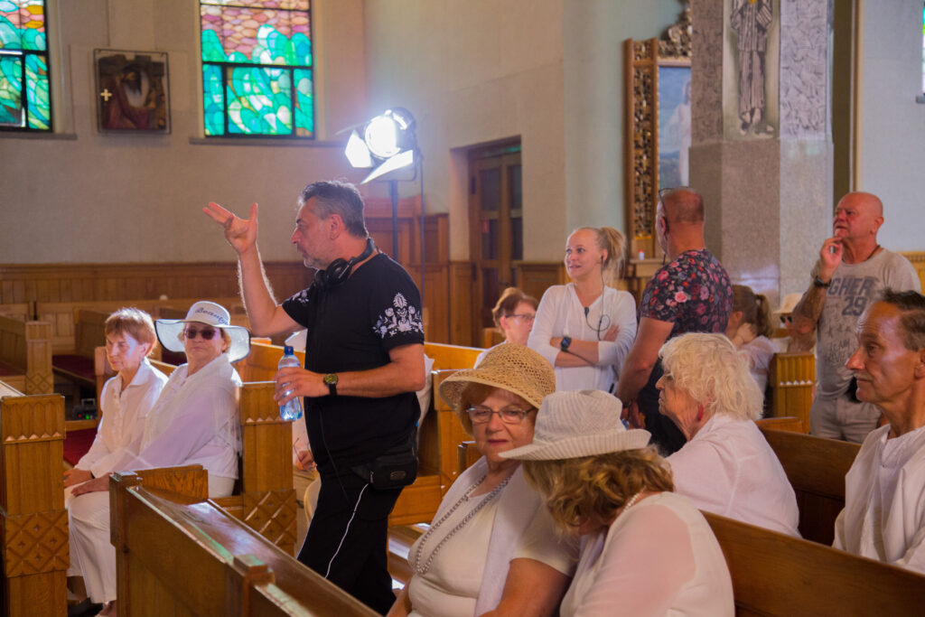 Wnętrze kościoła; w ławkach kościelnych siedzą goście weselni w parach. Wszyscy skupieni są na stojącym na środku reżyserze Arturze, który tłumaczy ruch kamery stojącej za nim ekipie filmowej.