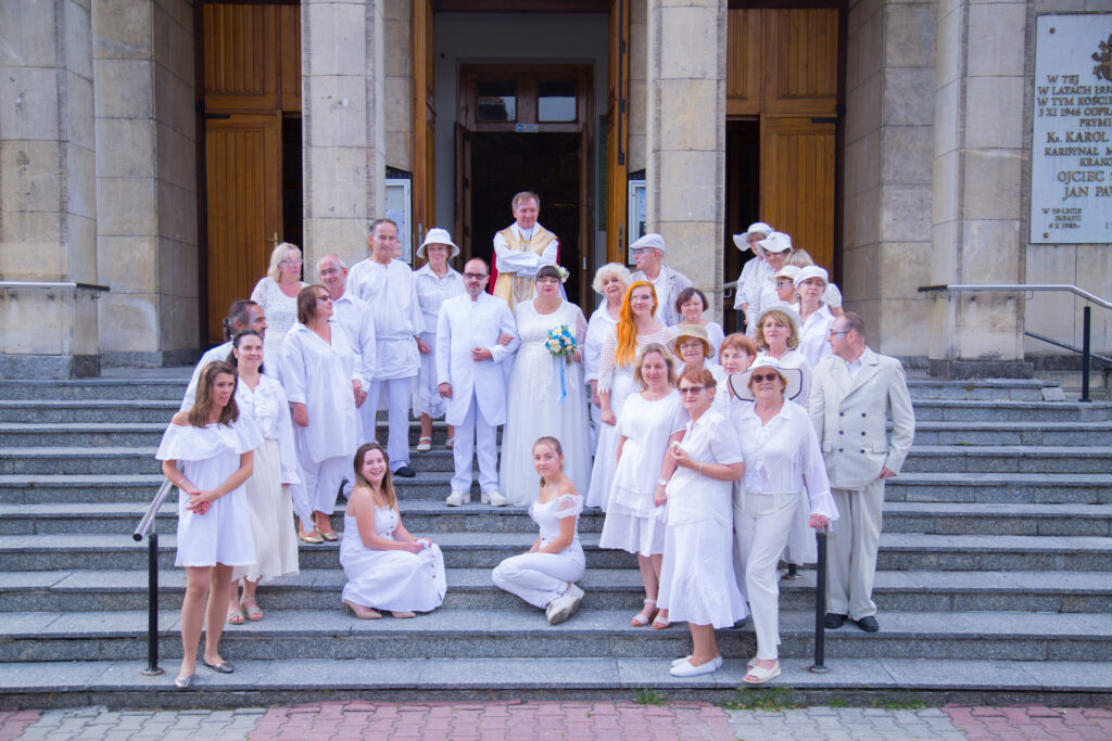 Zdjęcie grupowe; na schodach kościelnych stoi grupa ubranych na biało gości weselnych. W centrum grupy stoi para młoda, aktorzy Krzysiu i Monika. Za nimi, u szczytu schodów, stoi ksiądz. Wszyscy patrzą w obiektyw i się uśmiechają.