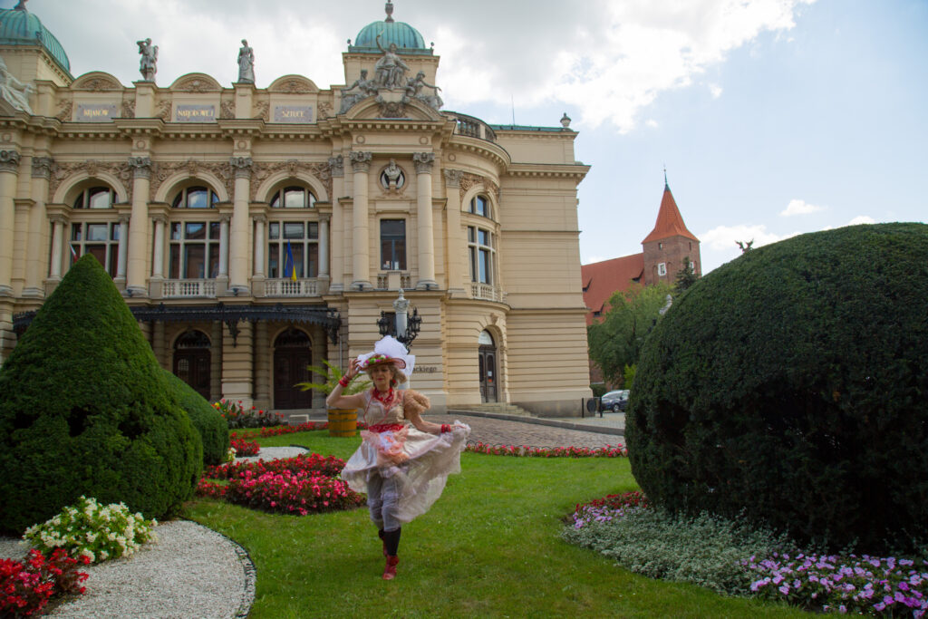 Teatr Słowackiego. W centrum zdjęcia aktorka Ela, przebrana w strój księżniczki ze spektaklu "Kota w butach", biegnie między kolorowymi klombami.