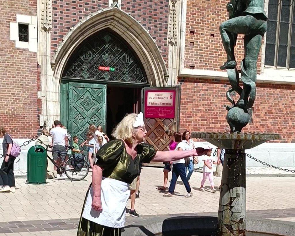 Plac Mariacki. W centrum zdjęcia fontanna z żakiem; aktorka Basia, przebrana w strój dwórki ze spektaklu  "Alicji w krainie czarów" obmywa rękę w spływającej wodzie. 