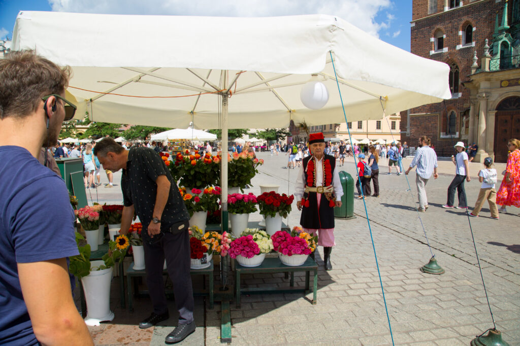 Rynek krakowski; w centrum zdjęcia jeden z kramów z kwiatami. Obok stoiska stoi aktor Janusz, przebrany w strój krakowiaka.