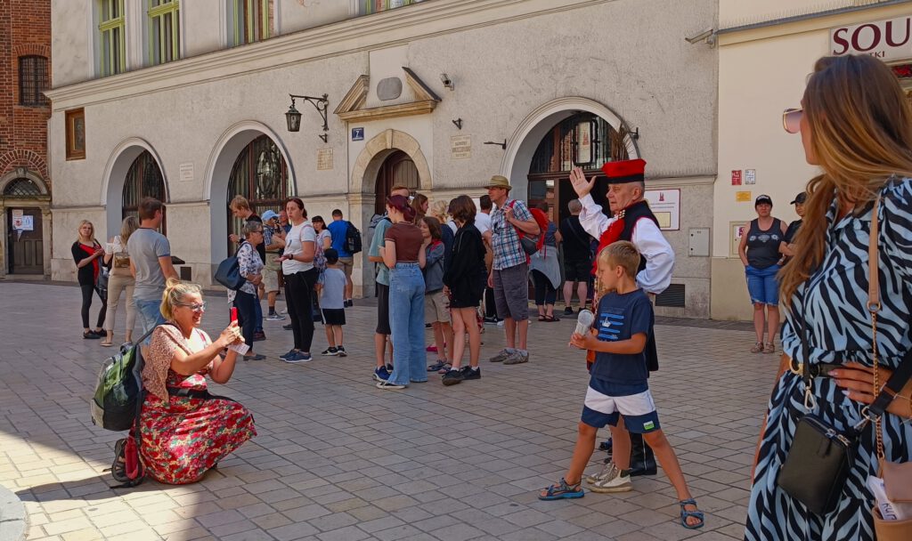 Plac mariacki; w tle tłum ludzi, w centrum matka robiąca zdjęcie dzieciom z aktorem Januszem, przebranym w strój krakowski.