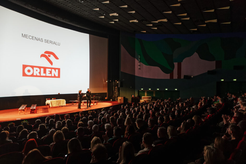 Zdjęcie sali kinowej wypełnionej gośćmi. Na scenie stoją producenci Danusia i Artur, na ekranie kinowym wyświetlony napis "Mecenas serialu: ORLEN"
