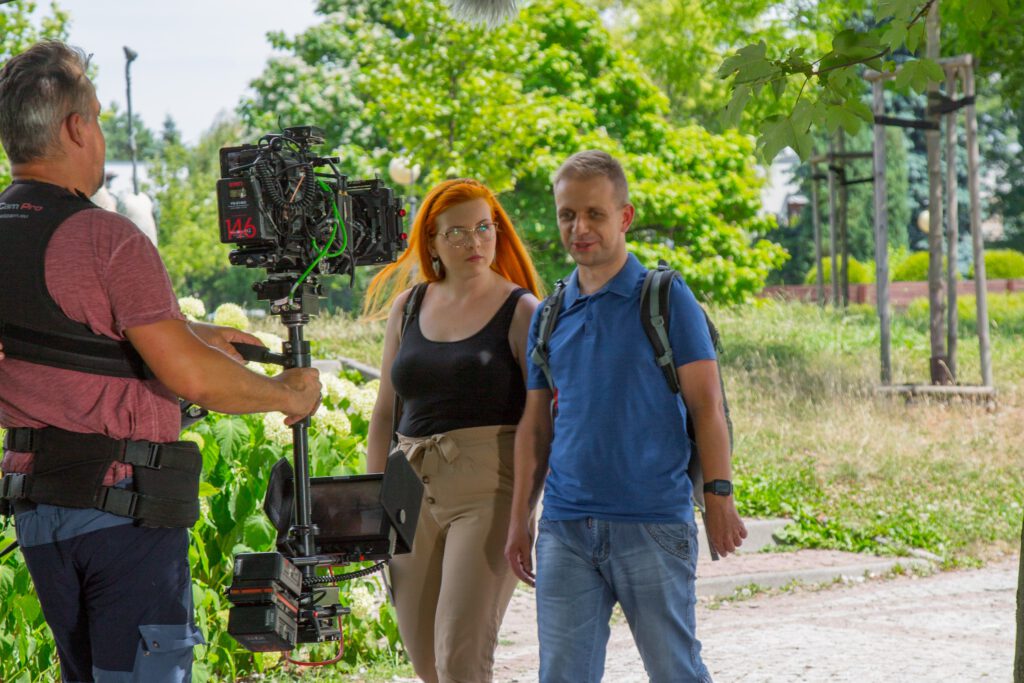 Aktorzy Aneta i Darek idą ścieżką w parku. Darek coś opowiada. Po lewej stronie widać operatora kamery nagrywającego scenę.