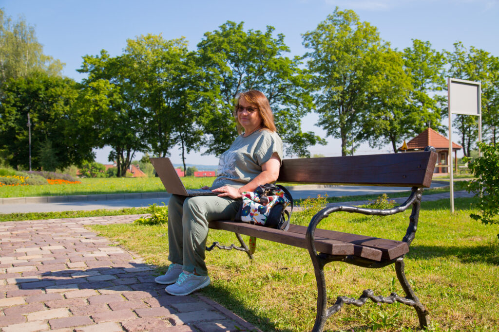 Ławka w parku Szpitala im. Babińskiego. Na ławce siedzi aktorka Danusia z laptopem na kolanach. Uśmiecha się do zdjęcia. 