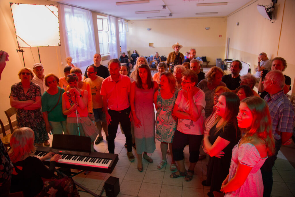 Grupa aktorów ITANa w czerwonym świetle ledowym stoi dookoła keyboardu i śpiewa. Na instrumencie gra akompaniatorka Monika.