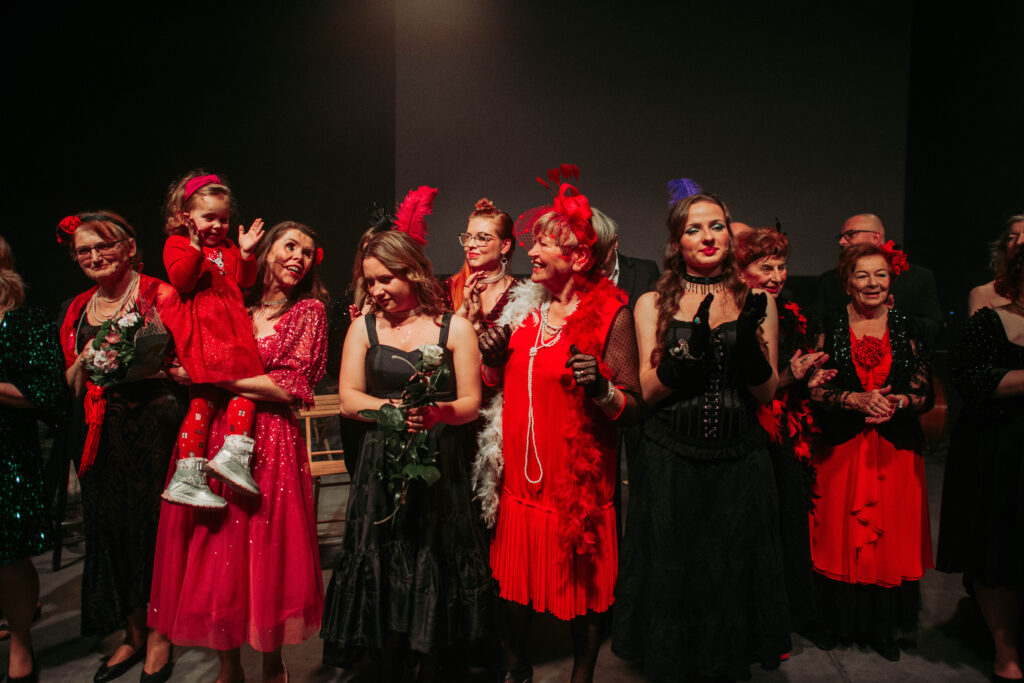 Zdjęcie kolorowe grupowe. Aktorzy w błyszczących czarno-czerwonych strojach stoją w grupie po ukłonach. Aktorka Gosia trzyma na rękach śmiejącą się siostrzenicę.