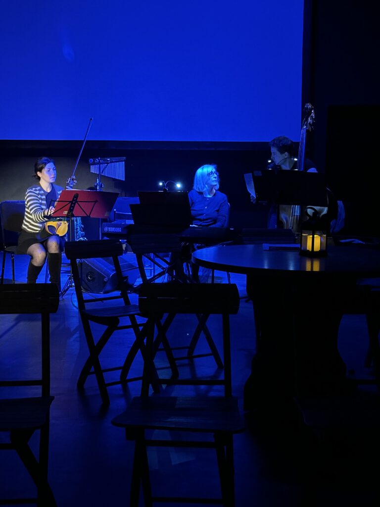 W centrum zdjęcia akompaniatorzy agnieszka, monika i krzysztof. Dookoła rozstawiona scenografia, stoliki i krzesła. SCena ośiwetlona na niebiesko. Muzycy rozmawiają.
