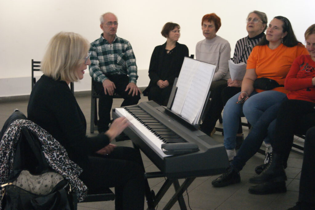 Akompaniatorka Monika gra na keyboardzie, dookoła niej zgromadzeni aktorzy śpiewają. Jedna osoba trzyma scenariusz.