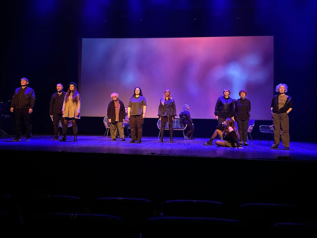 Zdjęcie ze spektaklu; scena w mocnym, niebieskim świetle. dziewięcioro aktorów stoi przodem do widowni, dwie aktorki siedzą tyłem - jedna na podłodze, druga na oparciu krzesła.  