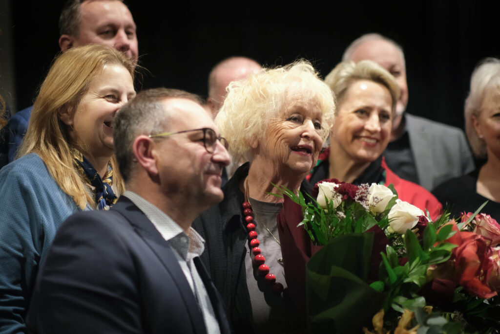 Gratulacje po premierze. W centrum zdjęcia aktorka Magda z bukietem biało-czerwonych róż. Dookoła elegancko ubrani ludzie. Wszyscy uśmiechnięci.